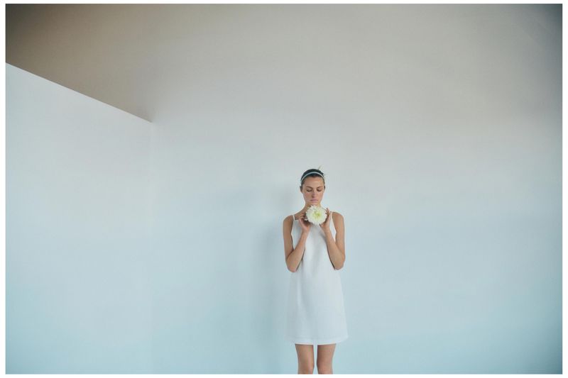 A young adult female in a white dress holds a white flower in an indoor setting with white walls and ceiling, creating a minimalist aesthetic.