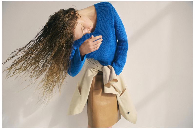Woman with wet curly hair wears a blue knit sweater and a beige jacket with a brown skirt in a studio setting.
