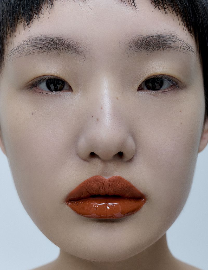 Close-up portrait of a young adult woman with dark eyes and short dark hair wearing shiny copper-colored lipstick on a white background.