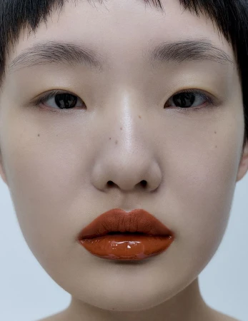 Close-up portrait of a young adult woman with dark eyes and short dark hair wearing shiny copper-colored lipstick on a white background.