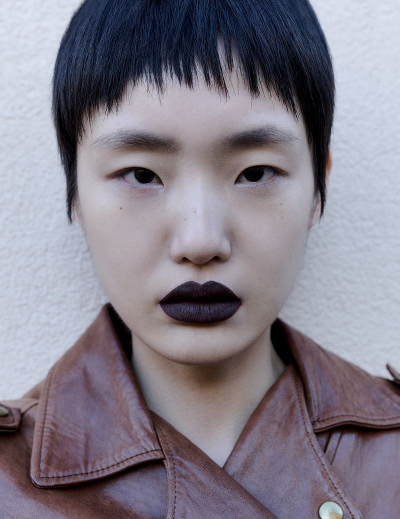 Close-up portrait of an adult female with short black hair, wearing a brown leather jacket and dark lipstick, against a neutral wall.