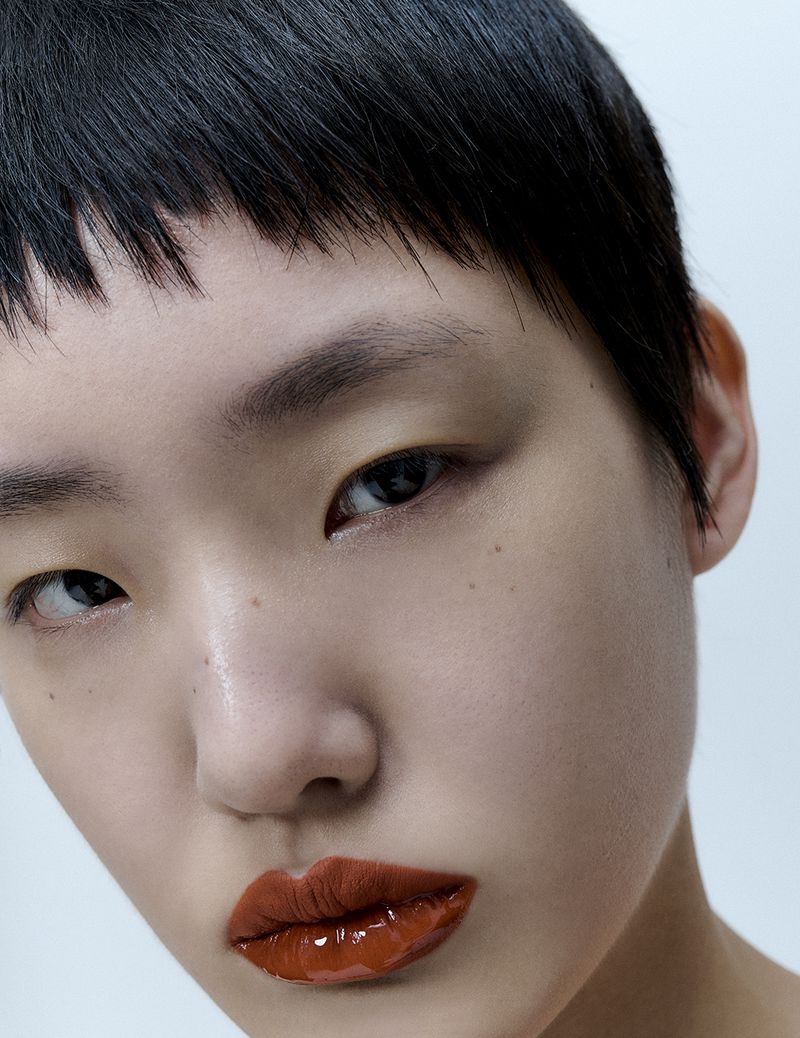 Close-up portrait of an Asian female with short black hair, glossy rust-colored lips, and light skin, set against a neutral white backdrop.