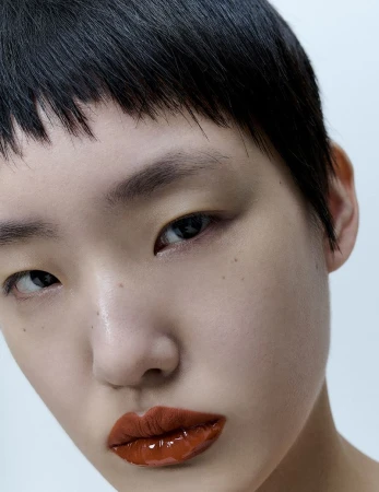 Close-up portrait of an Asian female with short black hair, glossy rust-colored lips, and light skin, set against a neutral white backdrop.