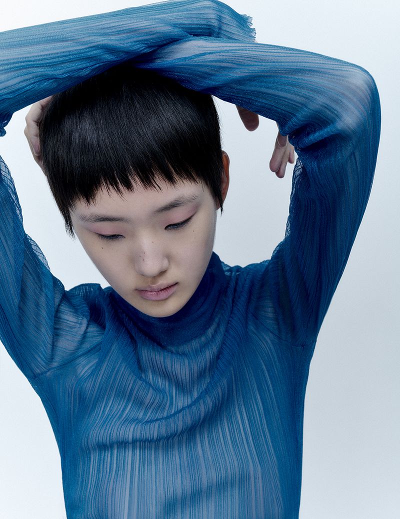 A young person poses indoors wearing a blue, textured, semi-sheer, long-sleeved top against a white background in a portrait-style image.