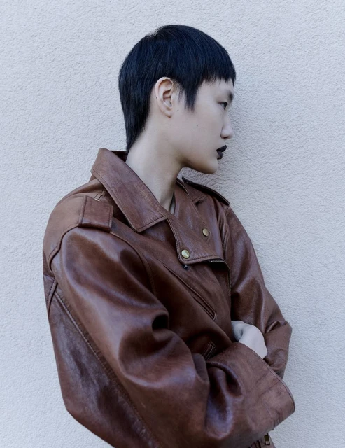 Side profile of a person with short dark hair wearing a brown leather jacket, posing in front of a neutral wall.