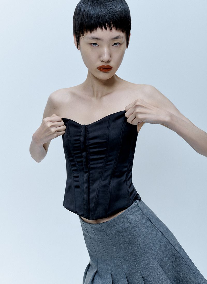 Model with short dark hair wearing a black strapless corset top and grey skirt against a pale grey background.
