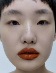 Close-up portrait of a woman with short dark hair, freckles, and glossy orange lipstick against a pale background.