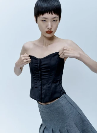 Model with short dark hair wearing a black strapless corset top and grey skirt against a pale grey background.