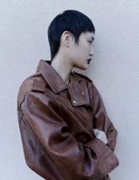 Profile portrait of a short-haired model wearing a brown leather jacket against a textured beige wall