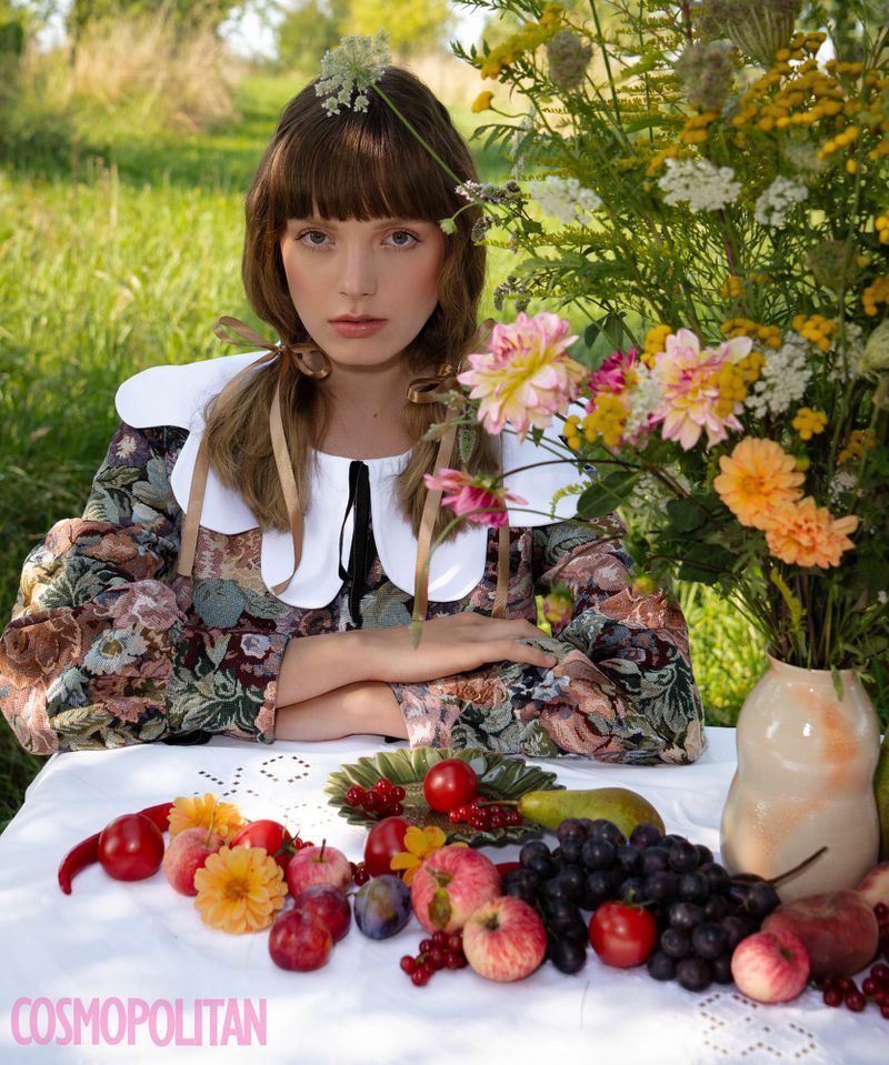 Young woman poses in floral dress at table laden with fruit and flowers, outdoors in a grassy field, editorial fashion shot.