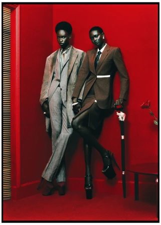 Two dark-skinned women pose indoors in fitted suits, one gray, one brown, against a red wall. One leans on a cane. Fashion editorial.