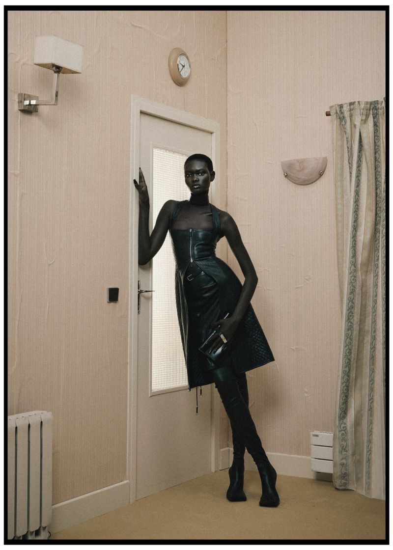 A slender Black woman poses in a black leather dress, boots, and purse against a beige interior, beside a white door and radiator.