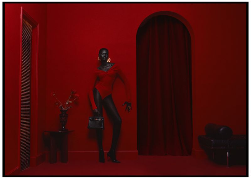 An adult woman with dark skin is posing in a red bodysuit and black tights, holding a black handbag against a red monochromatic interior.