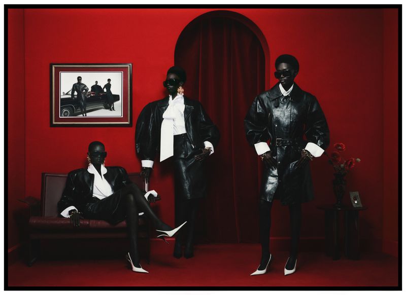 Three dark-skinned adult female fashion models in matching black-and-white outfits pose in a red interior with a framed photo.