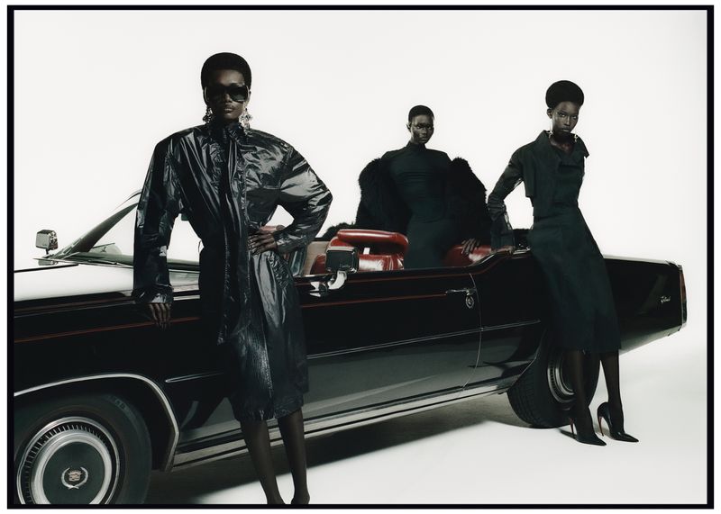 Three Black fashion models pose with a black classic Cadillac convertible in a photo studio setting, wearing sunglasses and fashionable dark clothing.