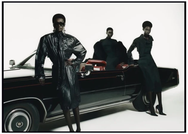 Three Black fashion models pose with a black classic Cadillac convertible in a photo studio setting, wearing sunglasses and fashionable dark clothing.