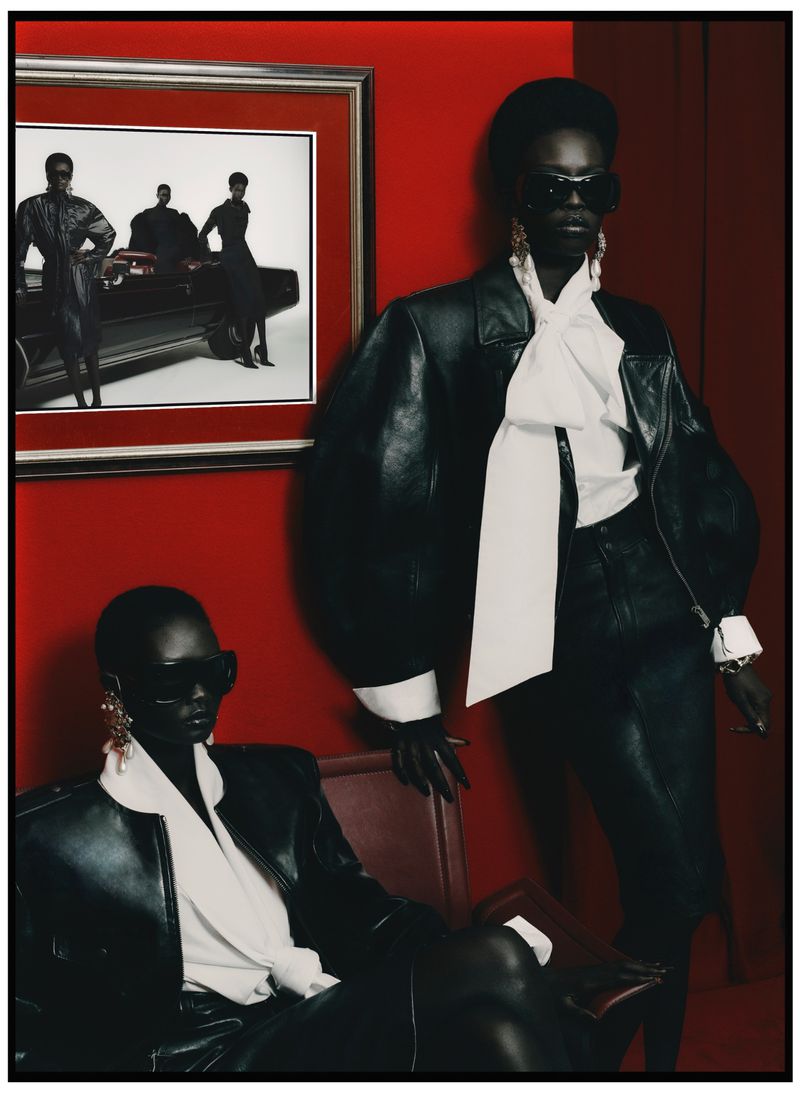 Two black women in leather jackets pose in a red room with a framed black-and-white photo, showcasing high-fashion editorial style.