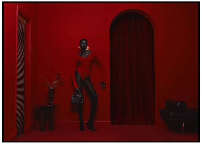 Model standing in a monochromatic red interior wearing a red bodysuit and holding a black handbag.