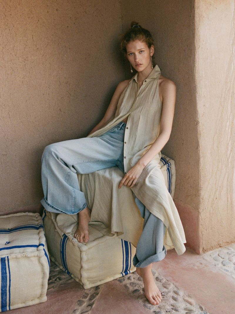 A fair-skinned woman poses in a beige sleeveless top and wide-leg jeans, seated on a striped cushion in a sunlit, stucco-walled setting.
