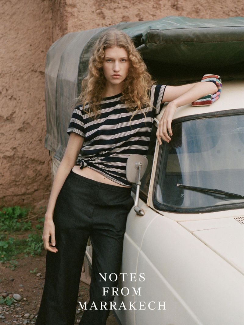 A woman with curly hair poses against a vintage car wearing a striped shirt and black pants for a fashion editorial shoot.
