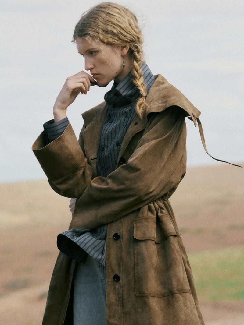 A fair-skinned woman with a blonde braid poses in a brown coat with a collared shirt in an outdoor setting with diffused lighting.