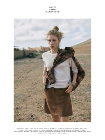 Model standing outdoors wearing a white t-shirt, brown skirt, and a plaid shirt draped over her shoulders