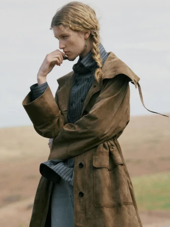 Model with a braided hairstyle wearing a brown coat and striped high-neck top outdoors against an open landscape.