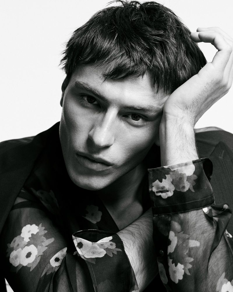 Portrait of a young adult male fashion model with dark hair, wearing a black blazer over a sheer floral-patterned shirt in a studio setting.