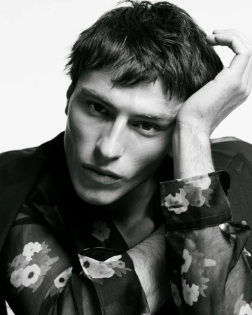 Portrait of a young adult male fashion model with dark hair, wearing a black blazer over a sheer floral-patterned shirt in a studio setting.