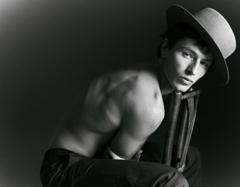 Monochrome portrait of a young man with bare upper body wearing a hat, seated on a wooden chair in a dark, shadowed studio.