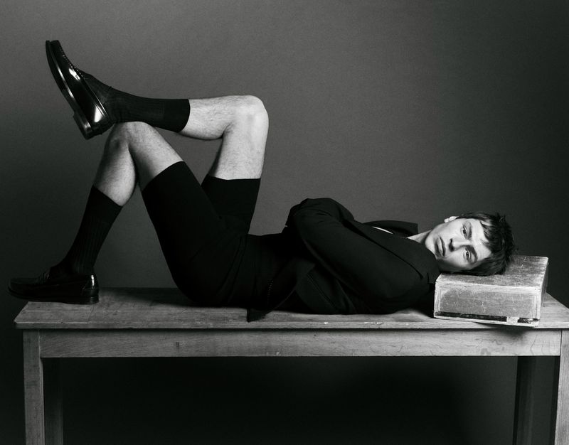 A man in black shorts, socks, jacket, and shoes is lying on a wooden table in a studio with a gray backdrop with his legs crossed.