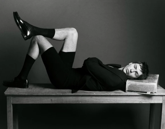 A man in black shorts, socks, jacket, and shoes is lying on a wooden table in a studio with a gray backdrop with his legs crossed.