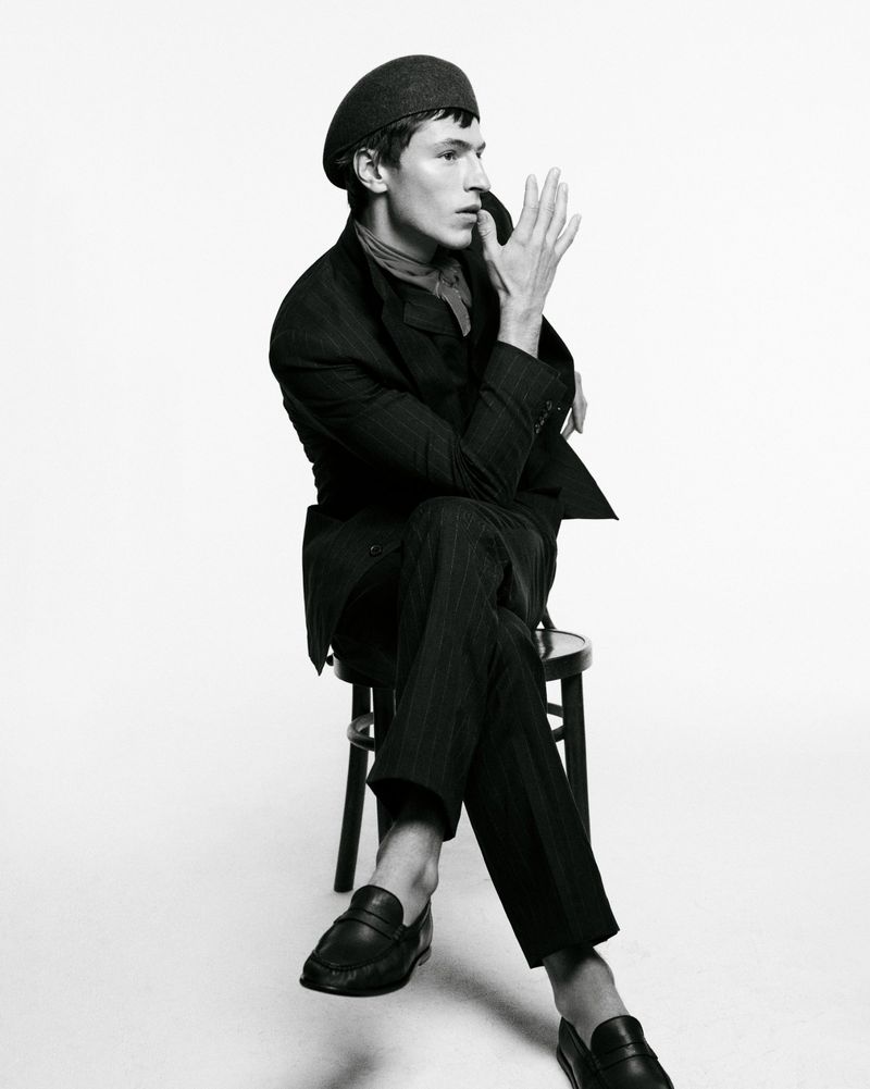 A young man with a dark beret and pinstripe suit sits on a black chair against a white background, one hand raised near his face.