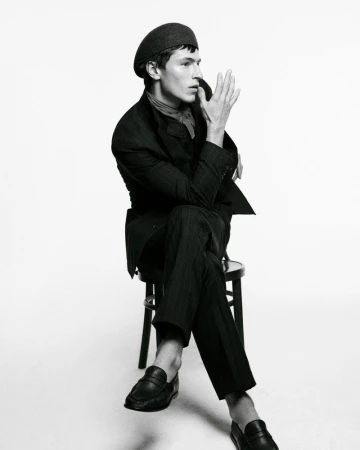A young man with a dark beret and pinstripe suit sits on a black chair against a white background, one hand raised near his face.