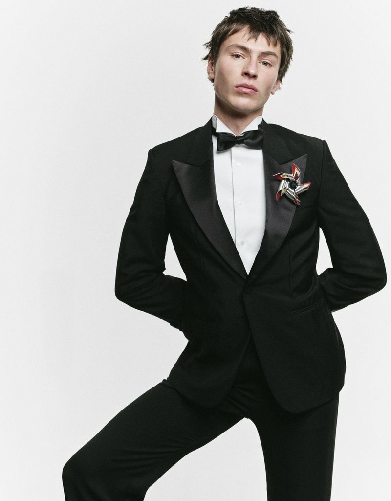A young adult male poses in a black tuxedo with a bowtie and decorative pin against a white background, looking confidently at the camera.