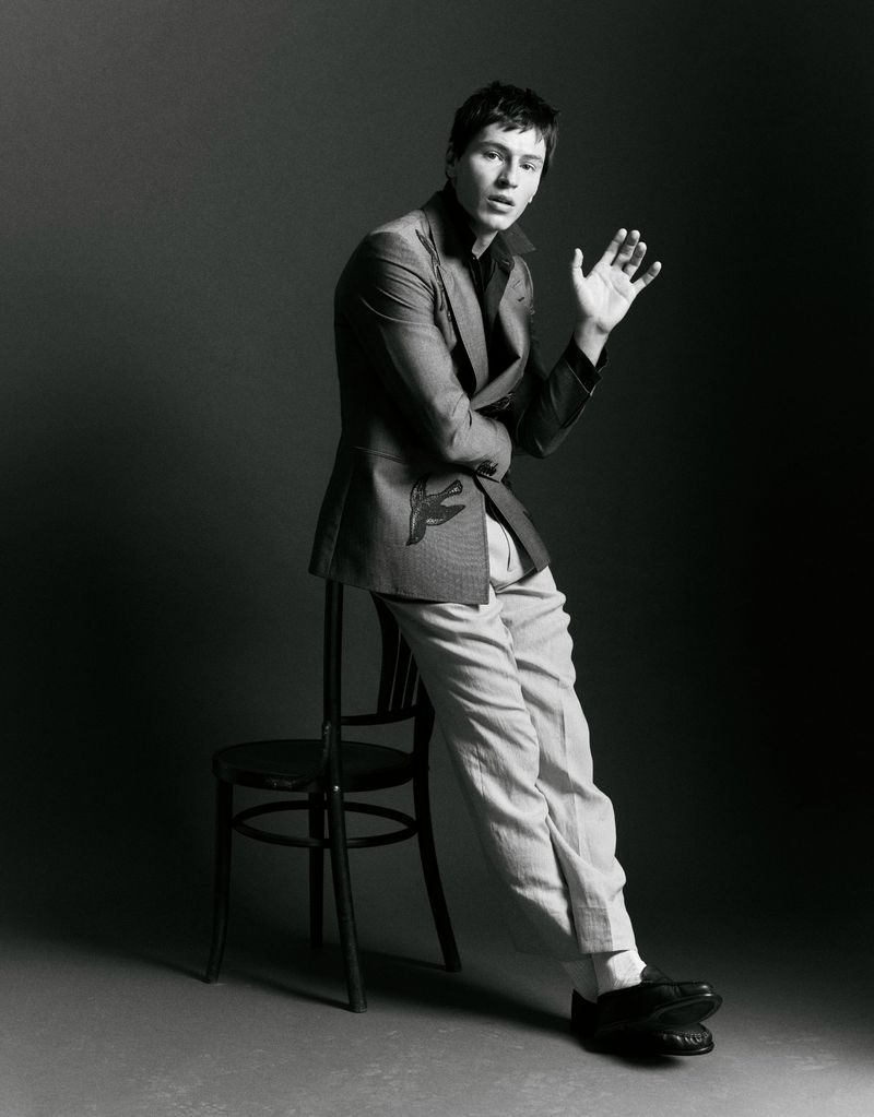 Portrait of a young adult male in a grey blazer and light trousers, leaning against a black chair in a photo studio setting.