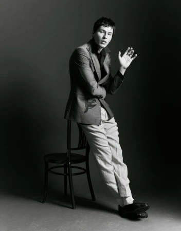 Portrait of a young adult male in a grey blazer and light trousers, leaning against a black chair in a photo studio setting.