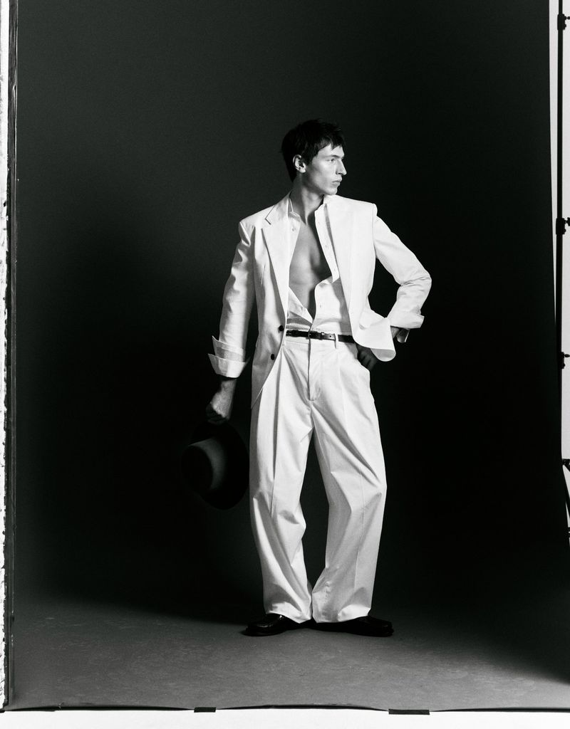 Man wearing a white suit and holding a dark hat against a grey seamless studio backdrop.