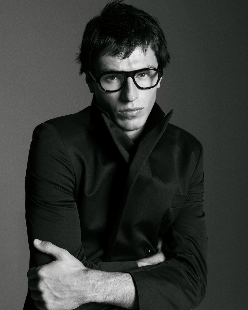 Black and white portrait of a man with glasses and a suit. He is indoors with a neutral background and arms crossed.