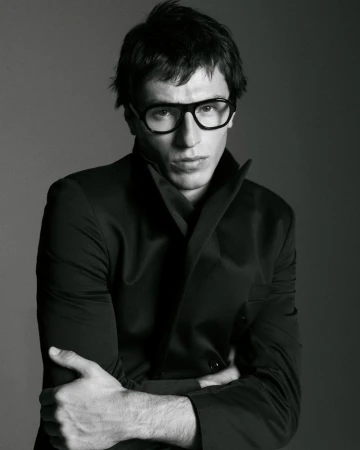 Black and white portrait of a man with glasses and a suit. He is indoors with a neutral background and arms crossed.