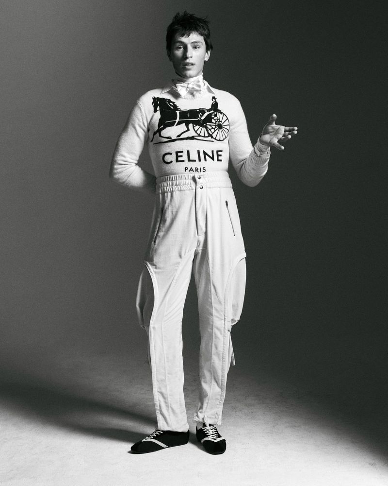 Monochrome portrait of a young adult man in a sweater with a horse-drawn carriage design, posing in a light grey photo studio.