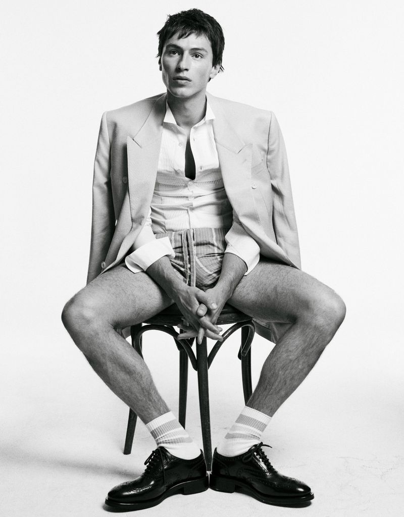 Man seated on a stool wearing a blazer draped over his shoulders, white shirt, and patterned shorts against a white backdrop.