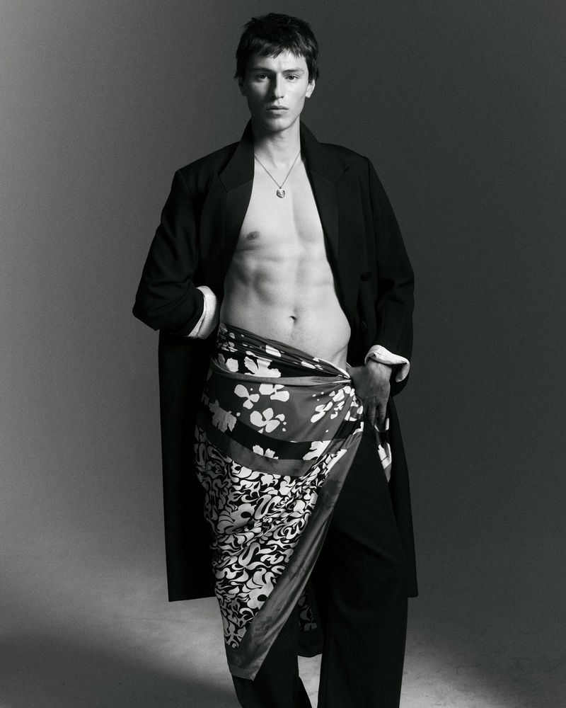 Young man with short dark hair standing shirtless wearing an open black coat and a floral patterned wrap skirt against a grey background.