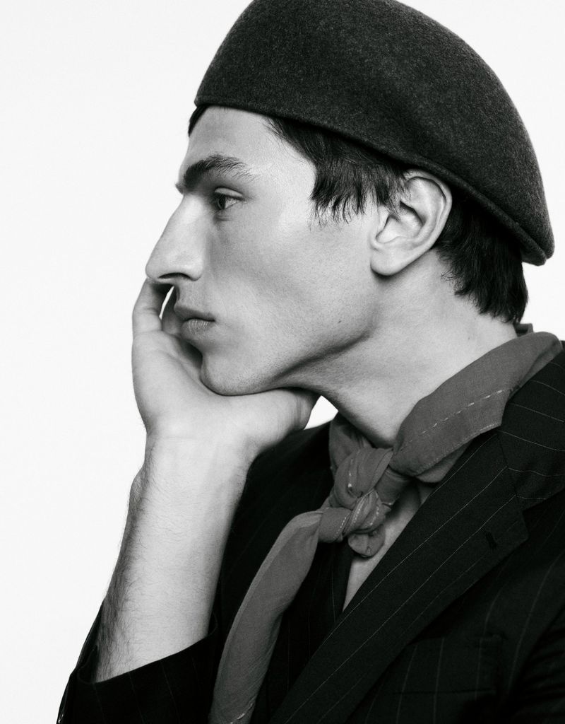 Black and white studio portrait of a young adult male with a beret, pinstripe jacket, and kerchief, resting his face on his hand.
