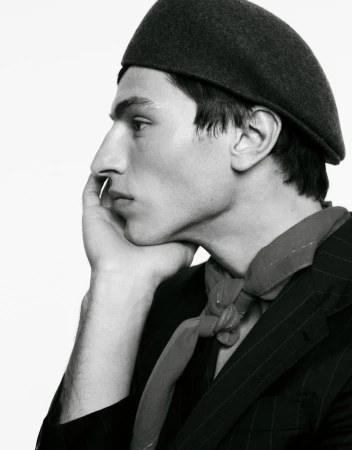 Black and white studio portrait of a young adult male with a beret, pinstripe jacket, and kerchief, resting his face on his hand.