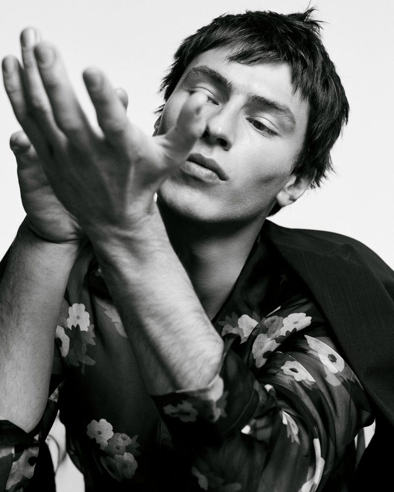 Close-up monochrome portrait of a male model holding hands up, wearing a floral shirt and dark jacket against a plain white background.