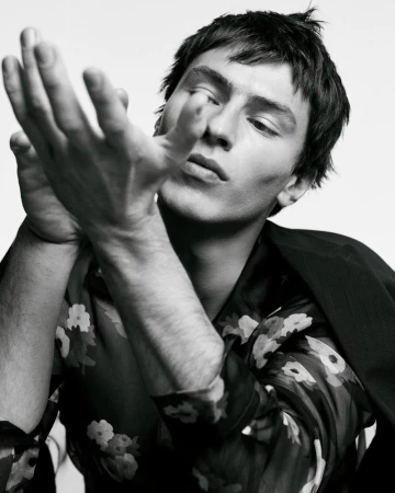 Close-up monochrome portrait of a male model holding hands up, wearing a floral shirt and dark jacket against a plain white background.