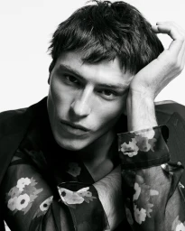 Close-up portrait of a male model in a black floral shirt with his hand resting against his head against a white background