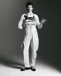 Monochrome portrait of a male model wearing a patterned sweater and white pants against a gradient studio backdrop.