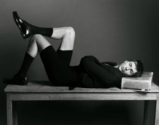 Monochrome fashion portrait of a male model reclining on a wooden table wearing dark shorts and loafers.